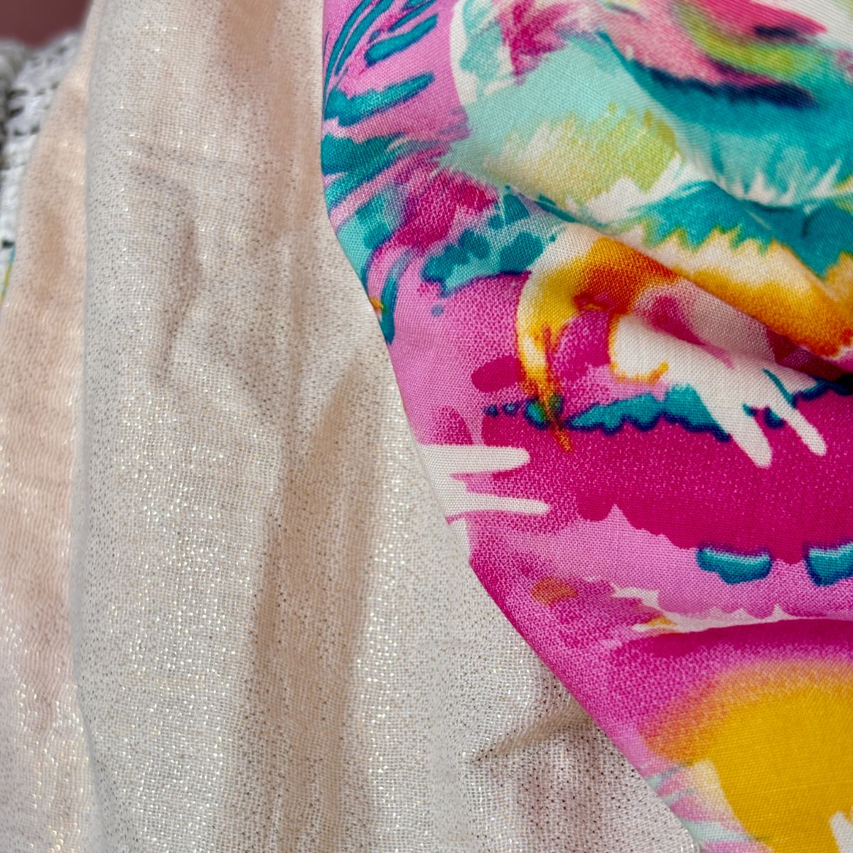 Gros plan sur les matières du chèche . Contraste entre la gaze de coton beige parsemée de fils dorés scintillants et le coton imprimé aux fleurs stylisées rose et turquoise.