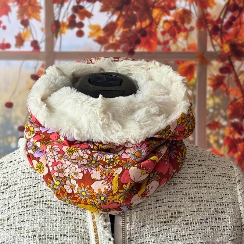 Maison Pivoine Un mannequin portant une veste blanche texturée présente le snood femme orange "Abélia" d'Echarpe, avec un motif floral et une doublure en fausse fourrure, les feuilles d'automne étant visibles à travers la fenêtre à l'arrière-plan. Fait main en Bretagne