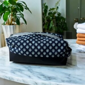 Maison Pivoine Une trousse de toilette homme bleue "Bouleau" en marine et noir repose sur un comptoir en marbre, complétée par des serviettes empilées et des plantes vertes en arrière-plan. Fait main en Bretagne