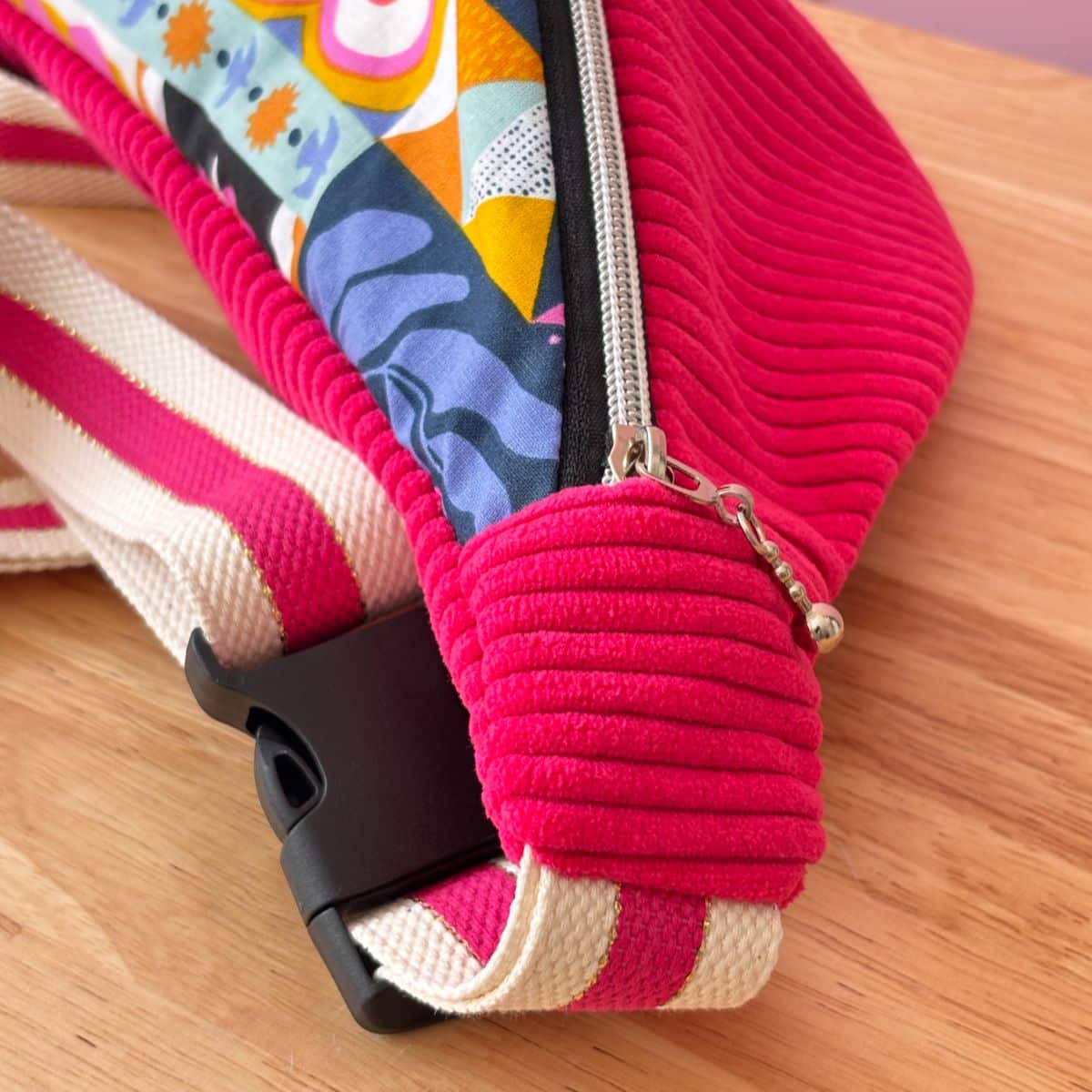 Zoom sur le côté du sac banane fuchsia : détail de la texture velours, de la sangle bicolore rose et blanc et de la boucle noire.
