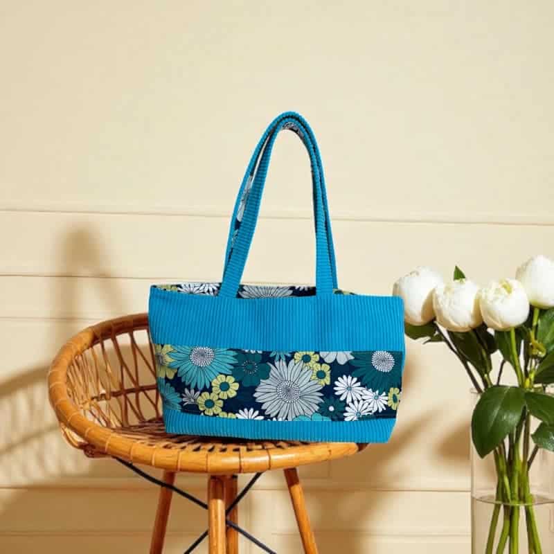 Sac cabas artisanal Maison Pivoine en velours côtelé turquoise et tissu fleuri vert et blanc, posé sur une chaise en rotin près de pivoines blanches.