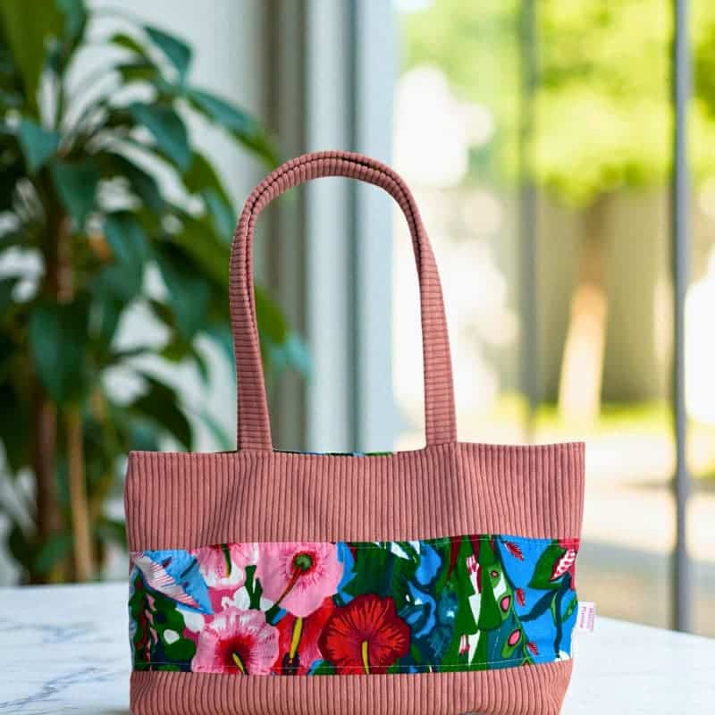 Sac cabas artisanal Maison Pivoine en velours côtelé rose avec bande fleurie multicolore, posé sur une table près d’une plante verte.