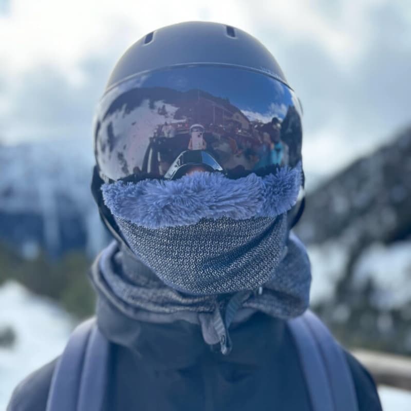 Personne équipée pour le ski portant un casque noir, un masque réfléchissant et un snood bleu doublé en fausse fourrure, dans un paysage de montagne enneigé.