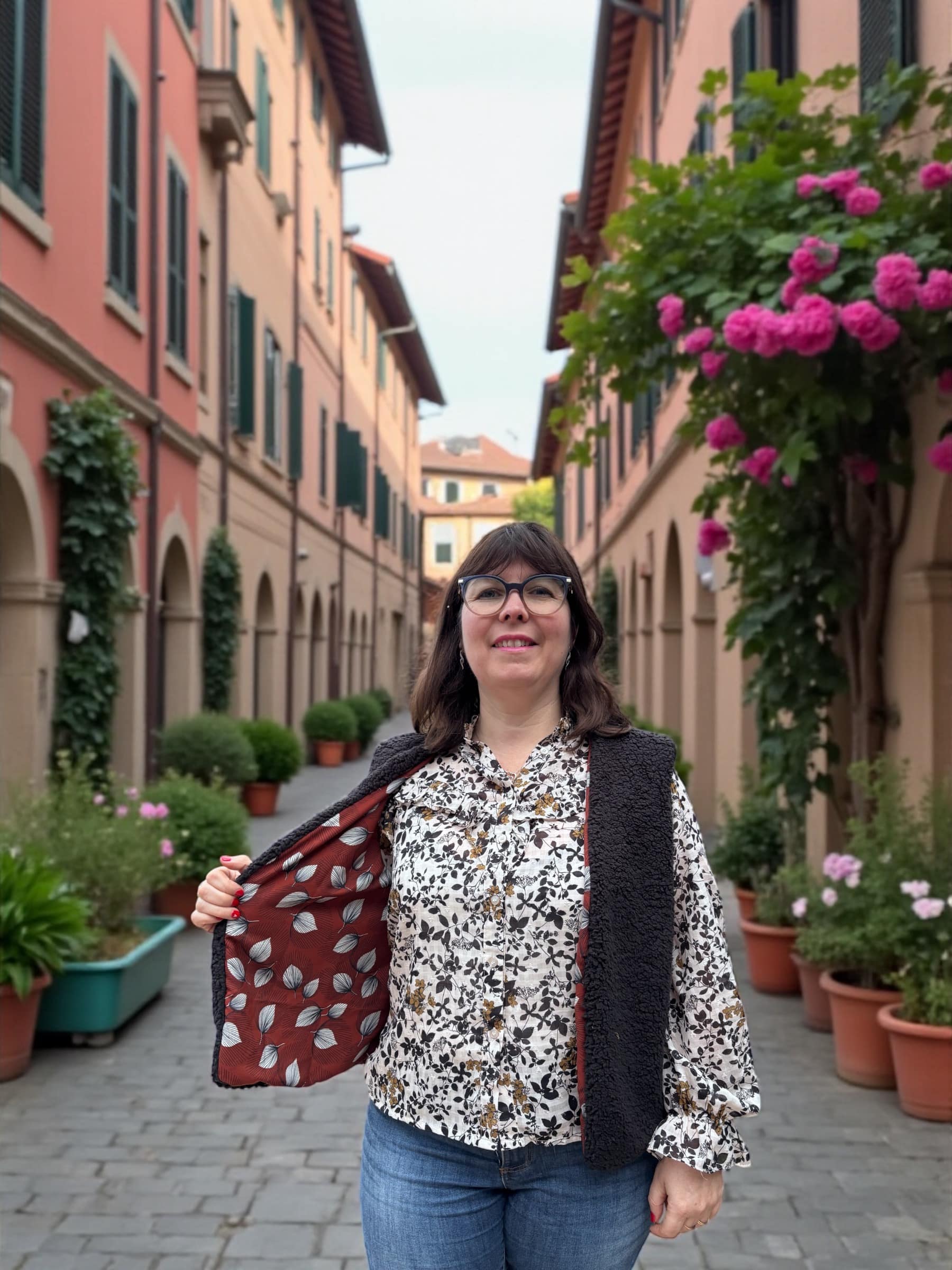 Femme portant un gilet de berger réversible, ouvrant le pan pour montrer la doublure viscose chocolat à motif feuilles, dans une ruelle avec des plantes en pots.