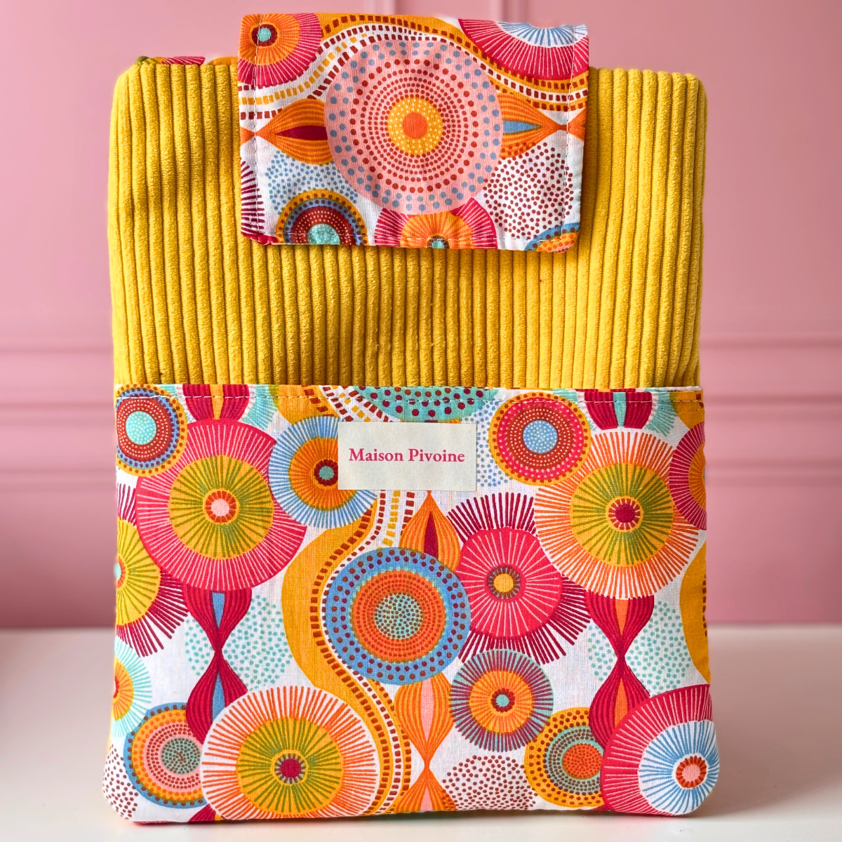 Pochette à livre de poche artisanale en velours côtelé jaune moutarde et grande poche avant en tissu fleuri aux motifs de rosaces colorées, avec étiquette Maison Pivoine.