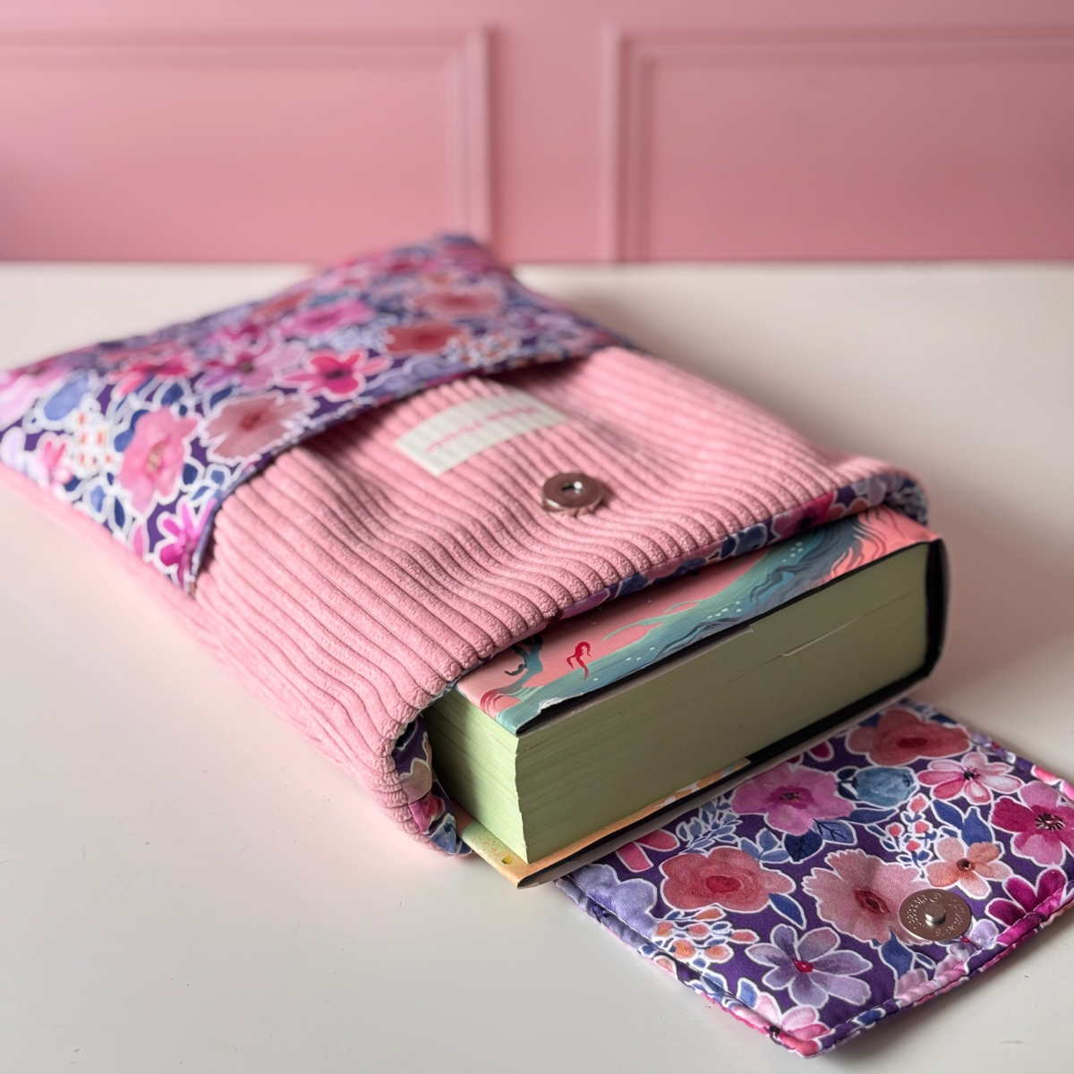 Pochette de protection pour roman en velours côtelé rose avec rabat ouvert laissant apparaître un livre, doublure et poche avant en tissu aux motifs floraux violets et roses.