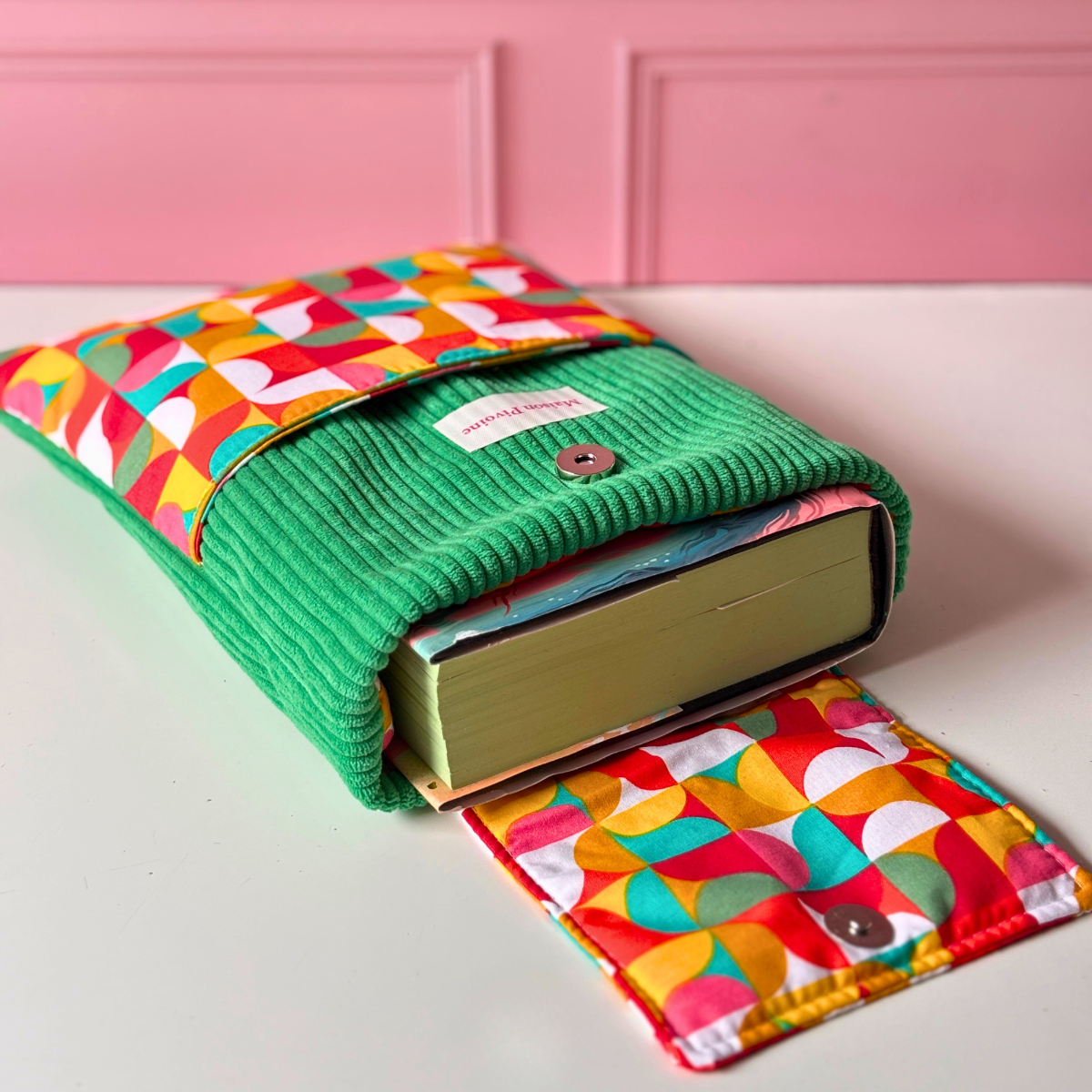 Pochette protectrice pour livre en velours côtelé vert sapin avec rabat aimanté ouvert montrant un gros roman, associée à un tissu aux motifs graphiques orange, jaune et vert.