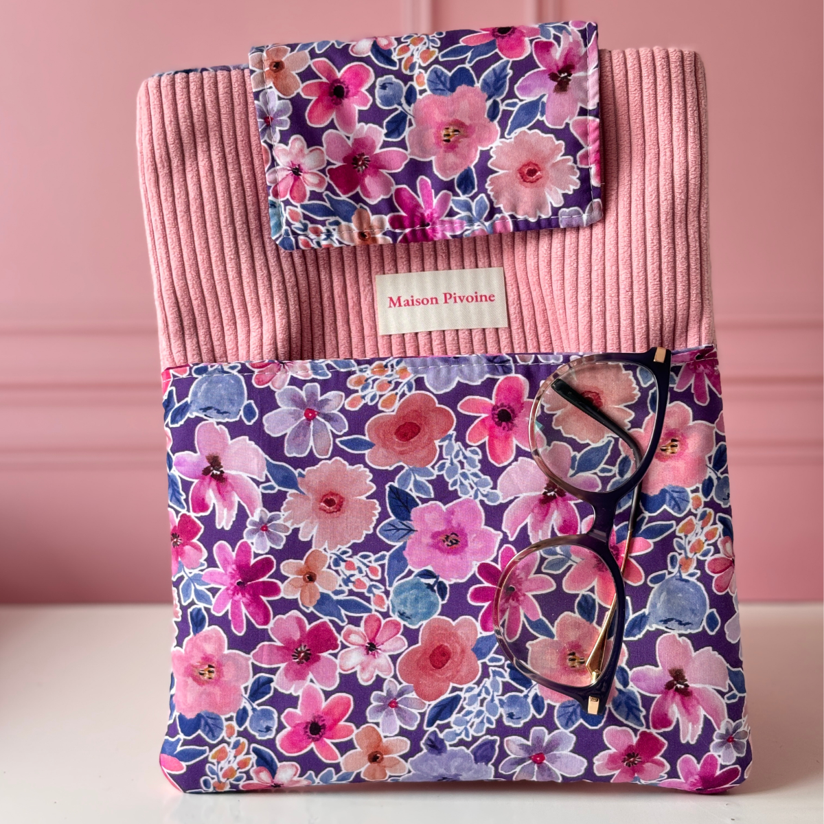 Vue de face d'une pochette à livre artisanale Maison Pivoine en velours côtelé rose et tissu liberty violet, avec une paire de lunettes posée sur la poche avant.