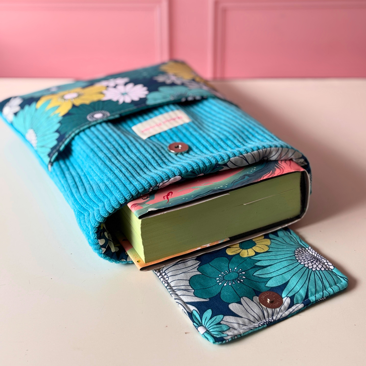 Pochette de protection pour livre de poche ou roman Maison Pivoine en velours côtelé turquoise et grande poche avant en tissu coton aux motifs de grosses fleurs rétro.